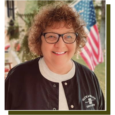 Portrait of smiling person with curly red hair wearing glasses and cardigan at dental office.