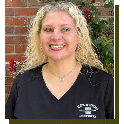 Professional headshot of smiling woman in black Glick & Woods Dentistry shirt against brick wall with flowers.