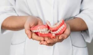 Dentist in white coat holding pink dentures in cupped hands against white background.