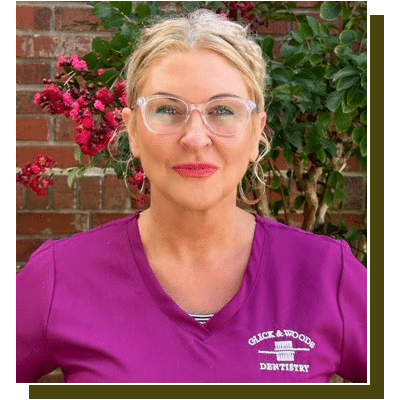 Professional woman wearing purple Glick & Woods Dentistry scrubs poses against brick wall with flowering vines.