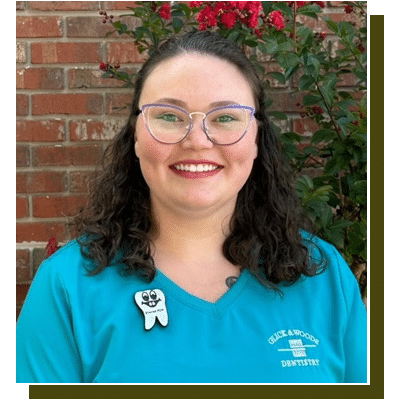 Smiling dental professional wearing blue scrubs and glasses against brick wall with ivy.