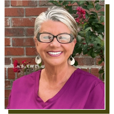Woman with short gray hair and glasses smiling at camera against brick wall with flowering vines.