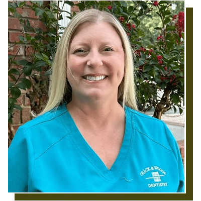 Woman in blue dental scrubs smiling at camera with brick wall and ivy background.
