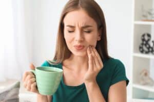 Woman holding green mug experiencing tooth pain, touching cheek in discomfort indoors.