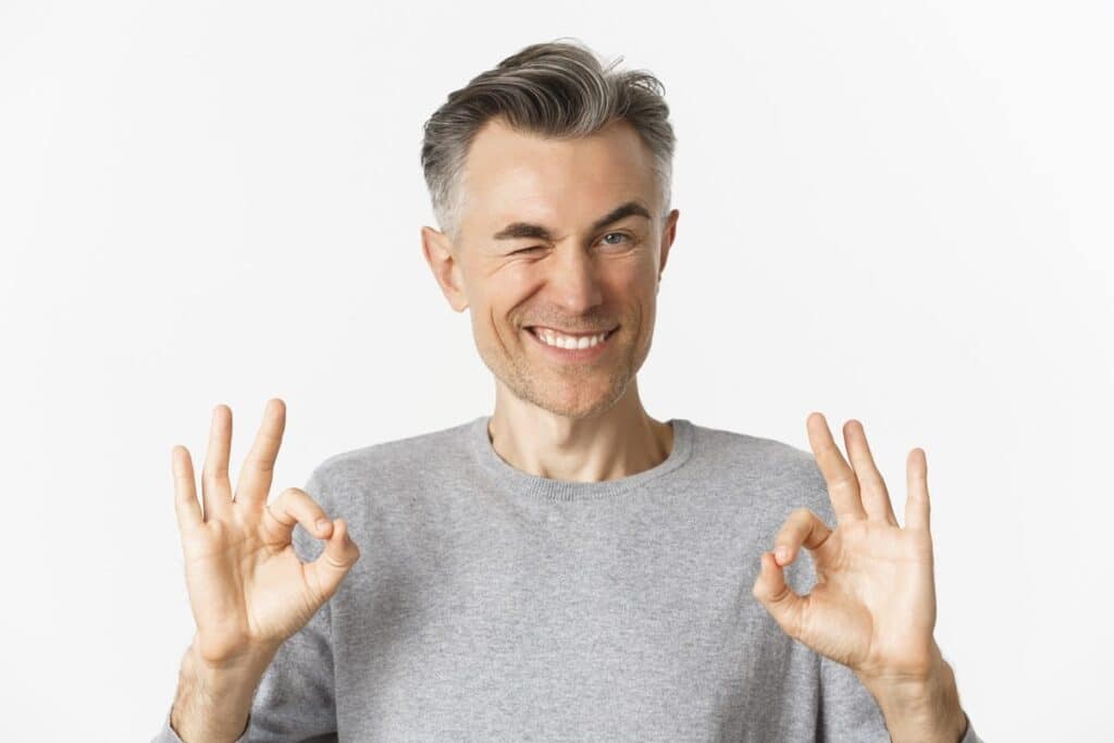 Middle-aged man in gray shirt making OK hand gestures, smiling against white background.