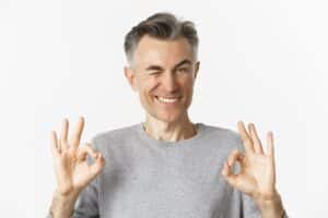 Middle-aged man in gray shirt making OK hand gestures, smiling against white background.