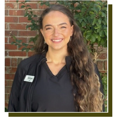 Smiling dental professional wearing dark scrubs with name badge against brick wall with ivy.