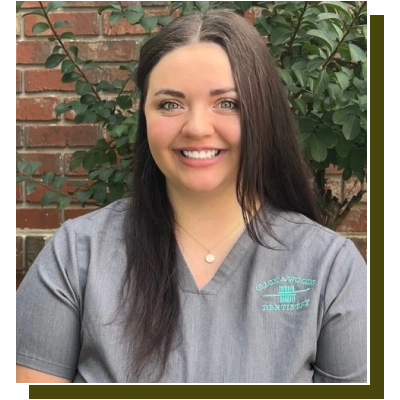 Professional female dental staff member smiling in scrubs against brick and ivy backdrop.