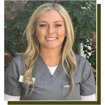 Professional female dental staff member smiling in scrubs against foliage background.