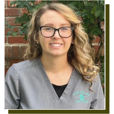 Young woman with glasses smiling at camera against brick wall background wearing dental scrubs.