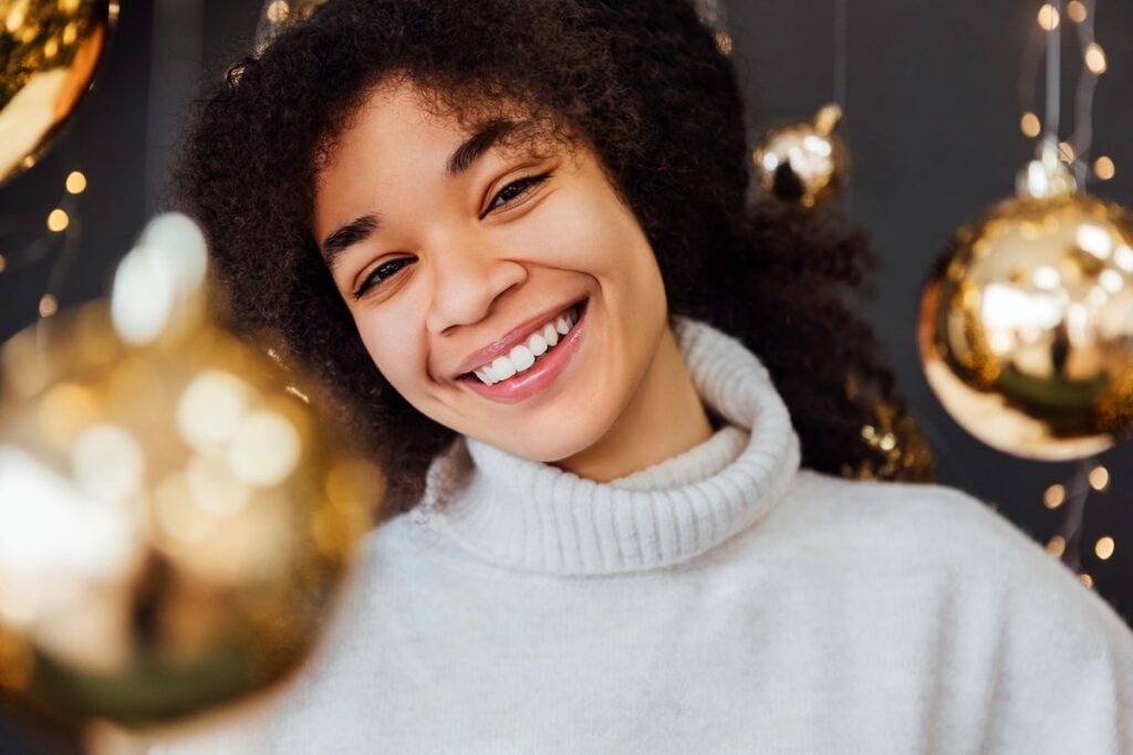 Smiling woman in white sweater against golden holiday decorations and bokeh lights background.