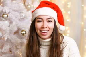 Woman wearing Santa hat smiling at camera with decorated Christmas tree and lights in background.