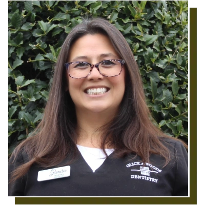 Professional headshot of female dental staff member wearing glasses and dark scrubs against ivy background.