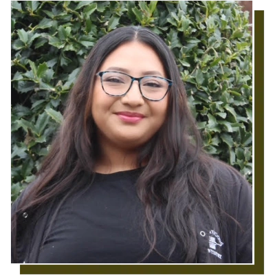 Young woman with glasses smiling outdoors against green ivy background, wearing black shirt.
