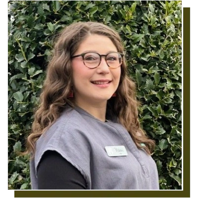 Young woman wearing glasses and dental office uniform smiling outdoors against green foliage.